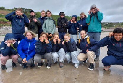 Group of young hikers on a sandy beach, some holding seaweed to their mouths in playful poses.