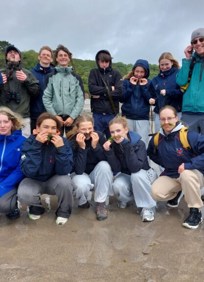 Group of young hikers on a sandy beach, some holding seaweed to their mouths in playful poses.