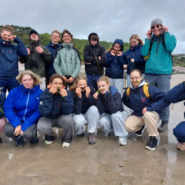 Group of young hikers on a sandy beach, some holding seaweed to their mouths in playful poses.