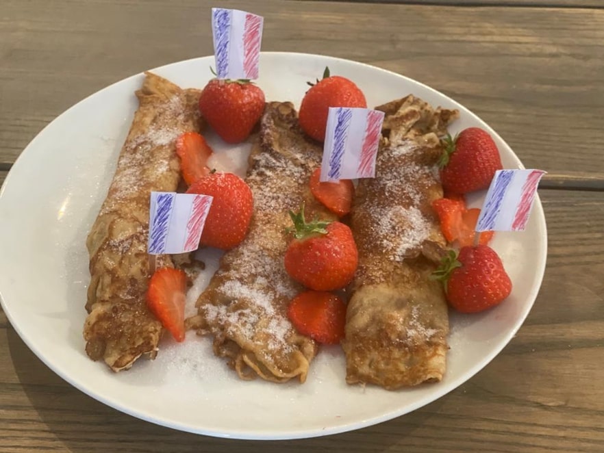Plate of crepes or pastries dusted with powdered sugar, surrounded by fresh strawberries, some halved, with small striped paper flags as decorations.