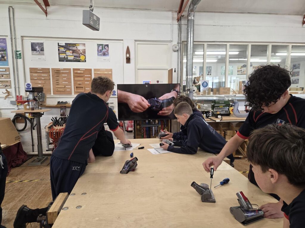Group of students at a woodworking workshop lean over a large workbench, using screwdrivers and tools on small devices while a wall-mounted screen shows a close-up of hands assembling a wristwatch.