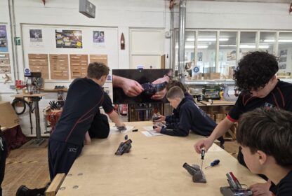 Group of students at a woodworking workshop lean over a large workbench, using screwdrivers and tools on small devices while a wall-mounted screen shows a close-up of hands assembling a wristwatch.
