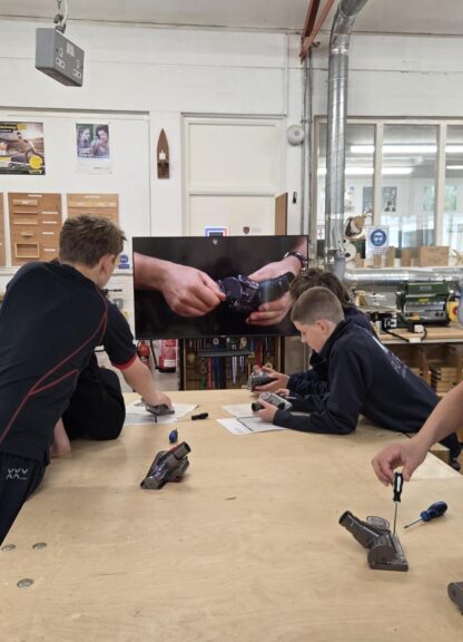 Group of students at a woodworking workshop lean over a large workbench, using screwdrivers and tools on small devices while a wall-mounted screen shows a close-up of hands assembling a wristwatch.