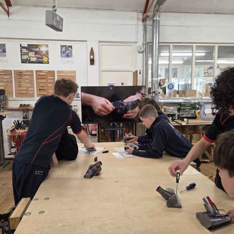 Group of students at a woodworking workshop lean over a large workbench, using screwdrivers and tools on small devices while a wall-mounted screen shows a close-up of hands assembling a wristwatch.