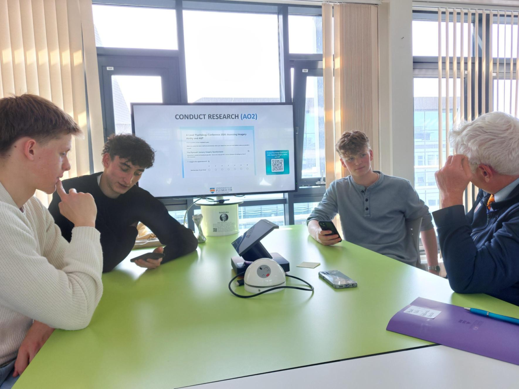 Group of students and a facilitator sit around a bright green table in a classroom, looking at their phones as a research presentation is shown on a screen in the background.