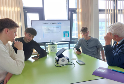 Group of students and a facilitator sit around a bright green table in a classroom, looking at their phones as a research presentation is shown on a screen in the background.