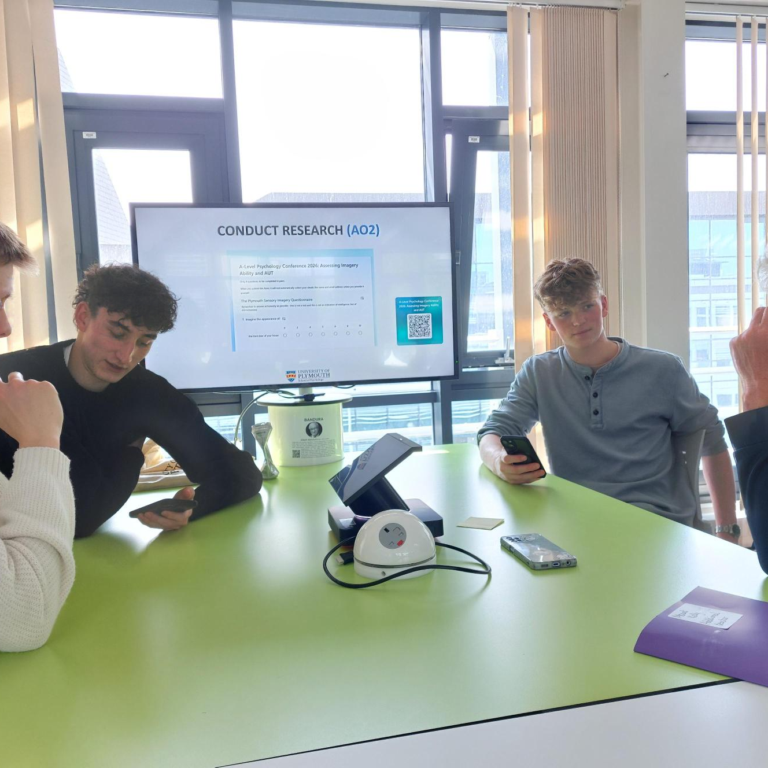 Group of students and a facilitator sit around a bright green table in a classroom, looking at their phones as a research presentation is shown on a screen in the background.