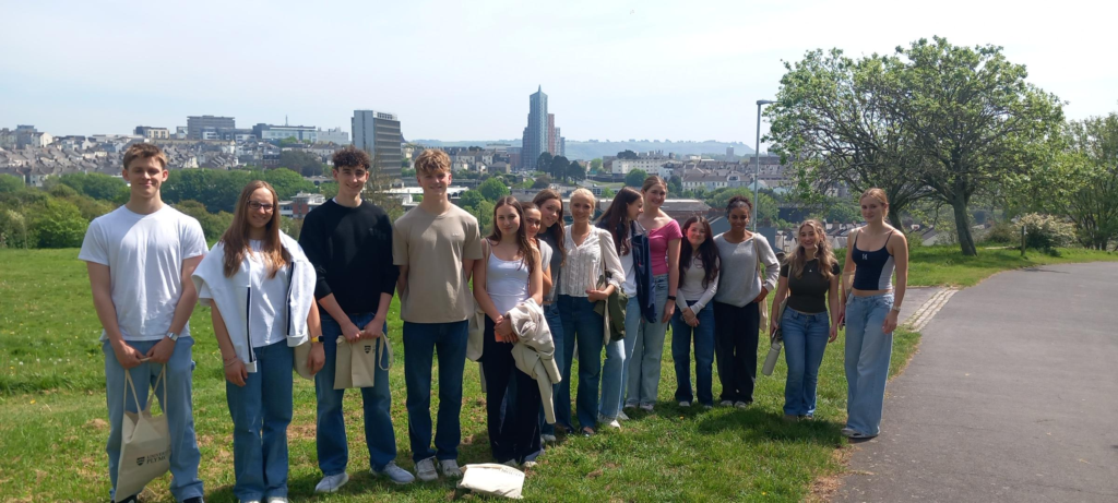 A group of young people standing in a line on a grassy hill with a cityscape skyline behind them, some holding bags and wearing casual clothes.
