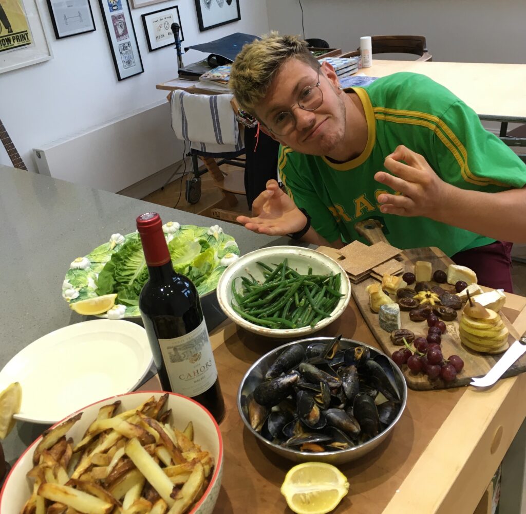 Person leaning toward the camera at a table crowded with food: bowls of fries, mussels, green beans, cheeses, grapes, lemons, and a bottle of red wine in foreground.
