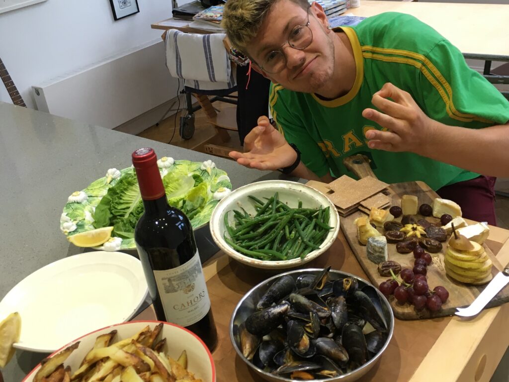 Person leaning toward the camera at a table crowded with food: bowls of fries, mussels, green beans, cheeses, grapes, lemons, and a bottle of red wine in foreground.
