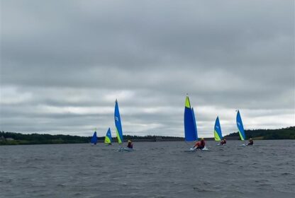 Group of small sailboats with bright blue and lime sails sailing on a gray, choppy lake under a cloudy sky, with people in life jackets.