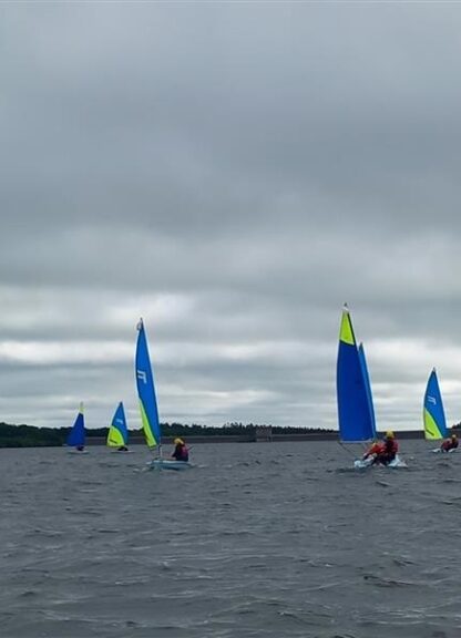 Group of small sailboats with bright blue and lime sails sailing on a gray, choppy lake under a cloudy sky, with people in life jackets.