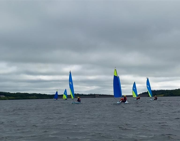 Group of small sailboats with bright blue and lime sails sailing on a gray, choppy lake under a cloudy sky, with people in life jackets.