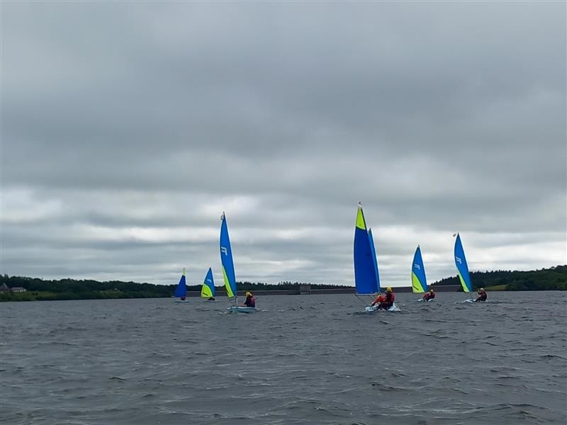 Group of small sailboats with bright blue and lime sails sailing on a gray, choppy lake under a cloudy sky, with people in life jackets.