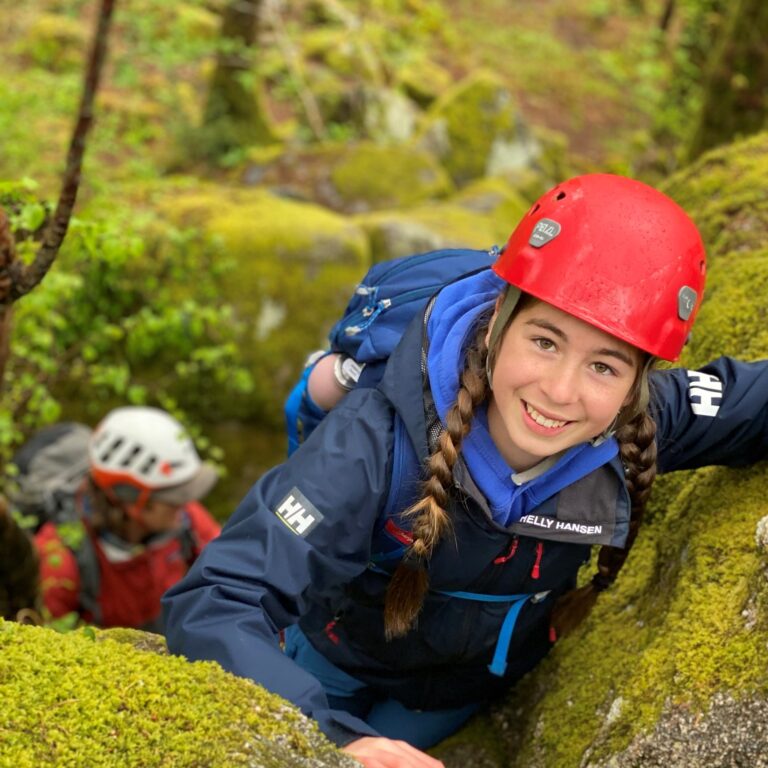 Young climber in a red helmet and blue jacket scales a mossy rock in a forest, with another climber visible in the background.