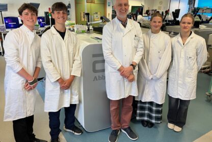 A group of five students in white lab coats smiling in a science lab with lab benches and equipment in the background.