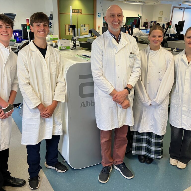 A group of five students in white lab coats smiling in a science lab with lab benches and equipment in the background.