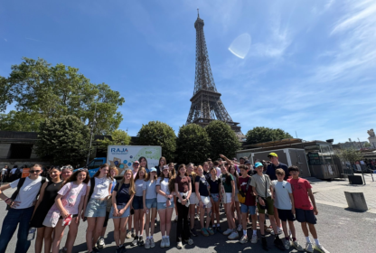 Group of students posing for a photo in front of the Eiffel Tower on a sunny day with blue sky.