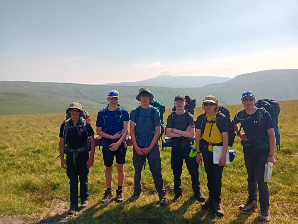 Six hikers with backpacks stand in a grassy hill meadow under a clear sky, posing for a group photo on a hiking trip.