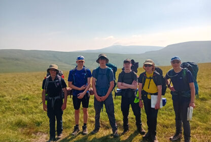 Six hikers with backpacks stand in a grassy hill meadow under a clear sky, posing for a group photo on a hiking trip.