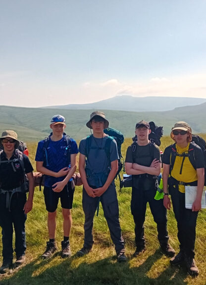 Six hikers with backpacks stand in a grassy hill meadow under a clear sky, posing for a group photo on a hiking trip.