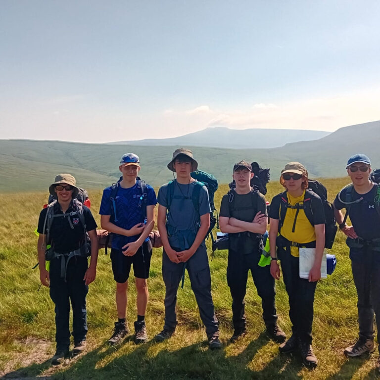 Six hikers with backpacks stand in a grassy hill meadow under a clear sky, posing for a group photo on a hiking trip.