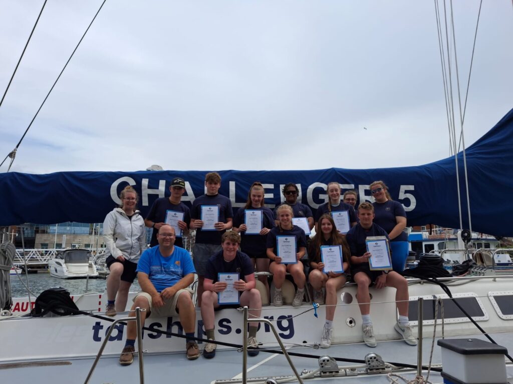 Group of young sailors on a sailboat holding certificates during a marina event, with a blue sail cover in the background.