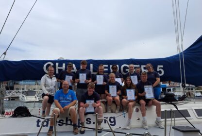 Group of young sailors on a sailboat holding certificates during a marina event, with a blue sail cover in the background.