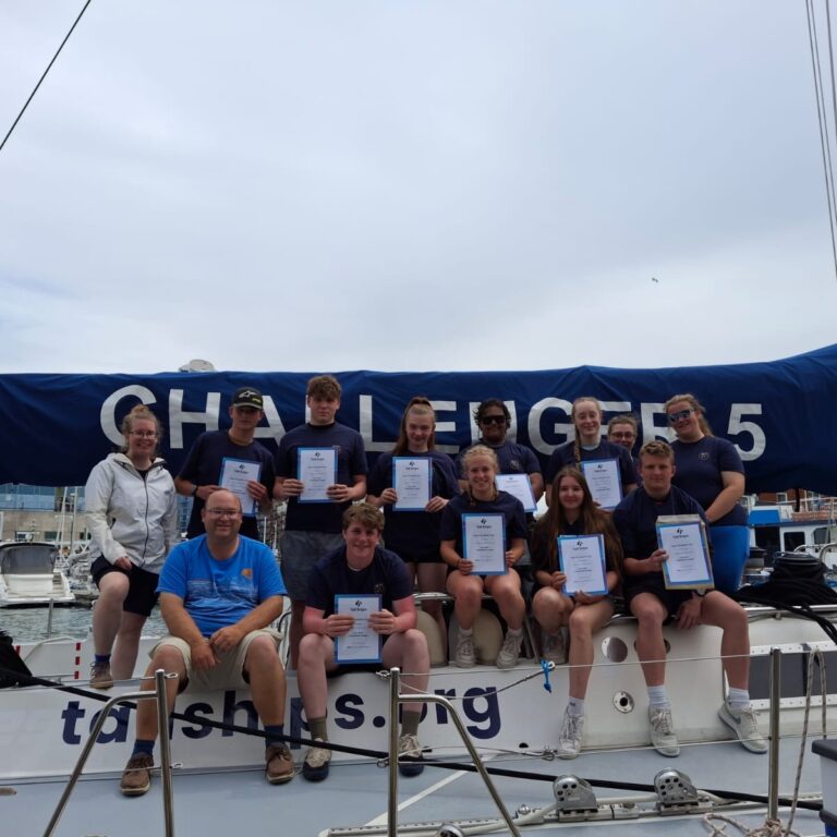 Group of young sailors on a sailboat holding certificates during a marina event, with a blue sail cover in the background.