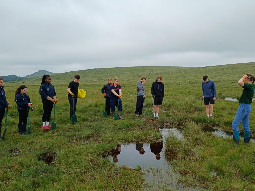 Group of young people with shovels lined up in a grassy field beside a shallow pond on a cloudy day.