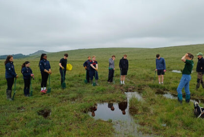 Group of young people with shovels lined up in a grassy field beside a shallow pond on a cloudy day.