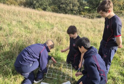 A group of students in navy uniforms kneel and examine a metal grid and maps in a tall grass field, with rolling countryside behind them.