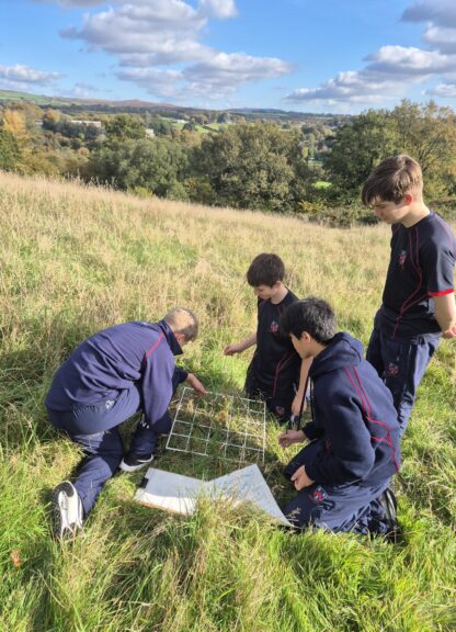 A group of students in navy uniforms kneel and examine a metal grid and maps in a tall grass field, with rolling countryside behind them.