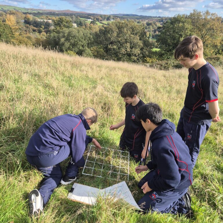 A group of students in navy uniforms kneel and examine a metal grid and maps in a tall grass field, with rolling countryside behind them.