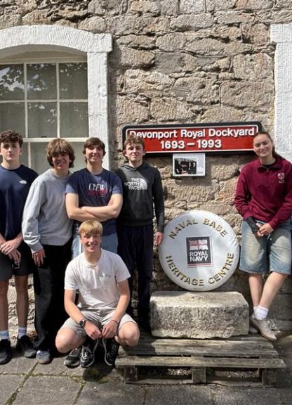 Nine teenagers standing in front of a stone building with a sign that reads 'Devonport Royal Dockyard 1693–1993' and a circular Royal Navy Heritage Centre emblem between them.