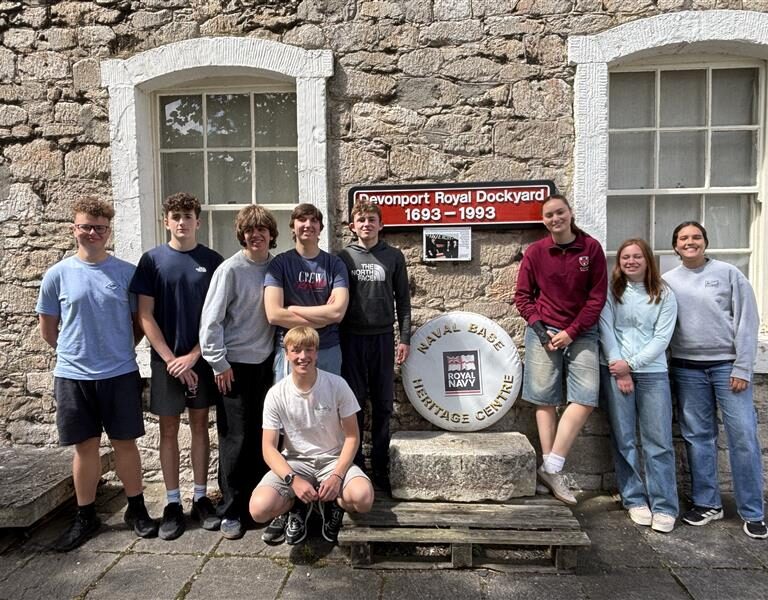 Nine teenagers standing in front of a stone building with a sign that reads 'Devonport Royal Dockyard 1693–1993' and a circular Royal Navy Heritage Centre emblem between them.